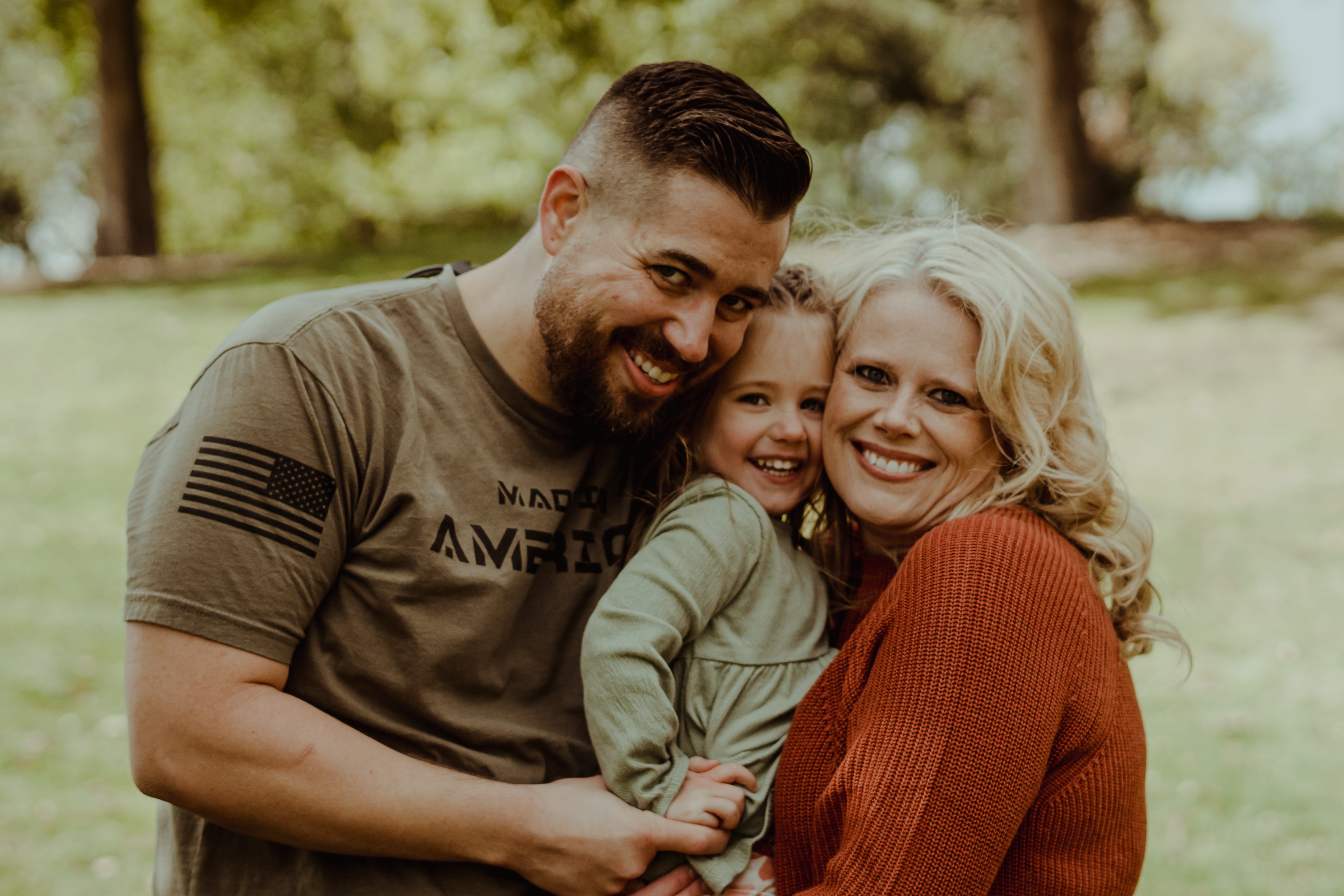 A happy family portrait featuring a man, a woman, and a young girl outdoors, smiling and embracing each other.