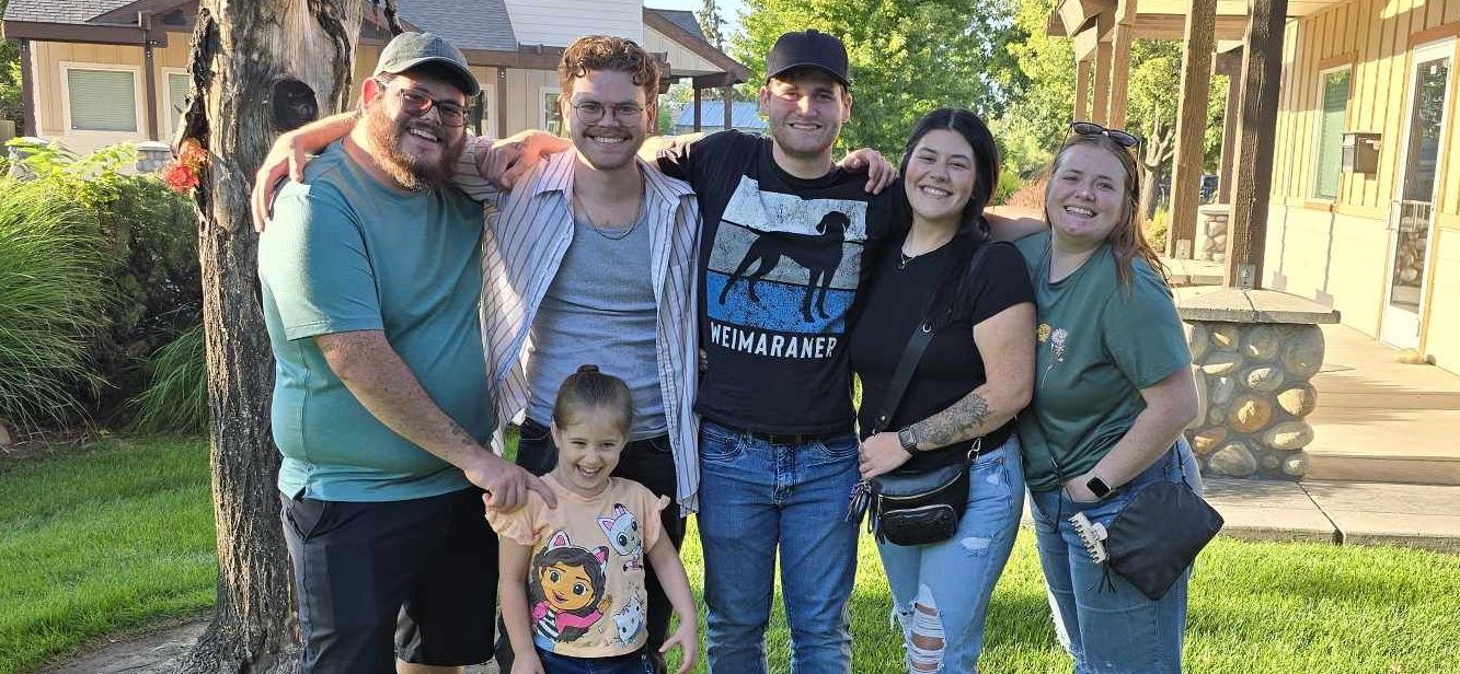 A group of six people posing outdoors, smiling and standing closely together. In the foreground, a young girl in a pink shirt is holding hands with an adult, while others are positioned around them. The background features greenery and a wooden structure.